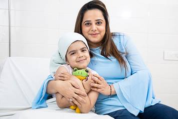 Mother embraces smiling daughter in hospital bed; child wears a headscarf and holds a stuffed frog plush toy.