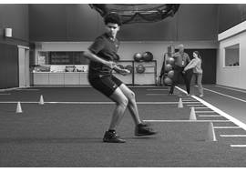 An athlete is running drills with cones in a large space. An athletic trainer and athlete are working together in the background.