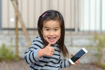 Adorable young Chinese girl smiling for a photo with a cell phone in one hand and a thumbs up in the other.