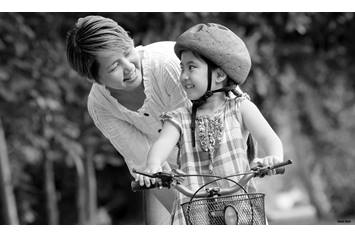 A smiling adult encourages a young girl learning to ride a bicycle in a park. The girl, wearing a helmet and a plaid dress, looks back at the adult with a joyful expression as they share a moment of support and connection.
