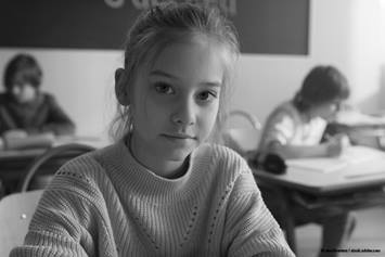 young girl in school wearing a turtleneck sweater with two students in the background sitting at their desks