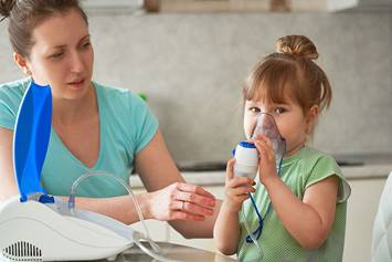 Little girl receives a breathing treatment using a nebulizer mask while her mother monitors the session at home.