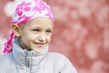 Young girl in a floral pink headscarf and gray jacket smiles brightly while standing outside against a blurred background.