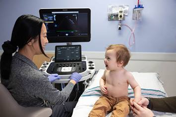 Smiling baby sits on a hospital bed holding caregiver's hand while a pediatric technician conducts an echocardiogram using an ultrasound machine.