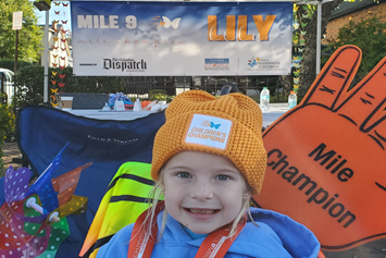 A smiling young child is wearing an orange hat. A "Mile 9 Lily" sign and a "Mile Champion" sign are behind her.