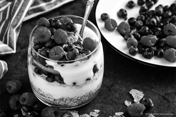 A glass filled with layers of granola, yogurt, blueberries, and raspberries sits on a dark surface with a spoon inside. Additional berries are scattered around, with a striped napkin and a plate of fruit in the background.