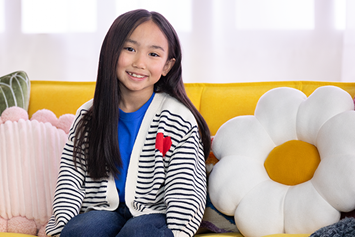 Smiling elementary aged girl sits on a mustard yellow couch with a daisy shaped pillow behind her.